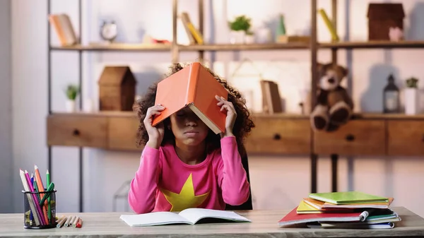 Gelangweiltes afrikanisch-amerikanisches Kind hält Buch auf dem Kopf, während es Hausaufgaben macht — Stockfoto