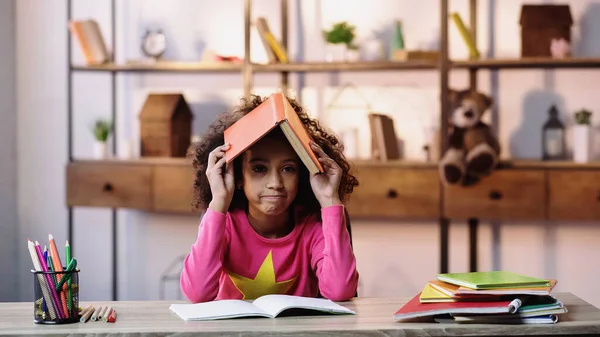 Enfant afro-américain mécontent tenant livre sur la tête tout en faisant des devoirs — Photo de stock