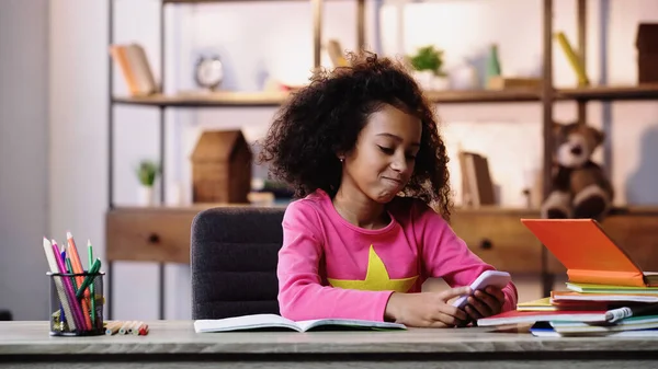 Enfant afro-américain joyeux en utilisant smartphone près des ordinateurs portables sur la table — Photo de stock