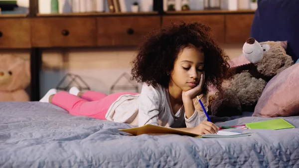 Gelangweilt afrikanisch-amerikanisches Kind macht Hausaufgaben, während es im Bett liegt — Stockfoto