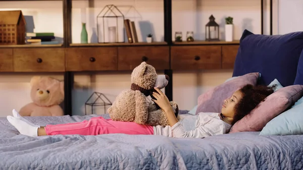 Vista lateral de rizado afroamericano chica acostada en la cama y mirando osito de peluche - foto de stock