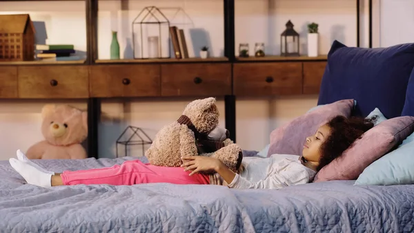 Vista lateral del niño afroamericano rizado acostado en la cama y mirando al oso de peluche - foto de stock