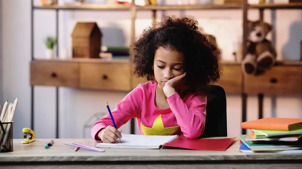 Entediado afro-americano menina escrevendo no notebook enquanto fazendo lição de casa — Fotografia de Stock