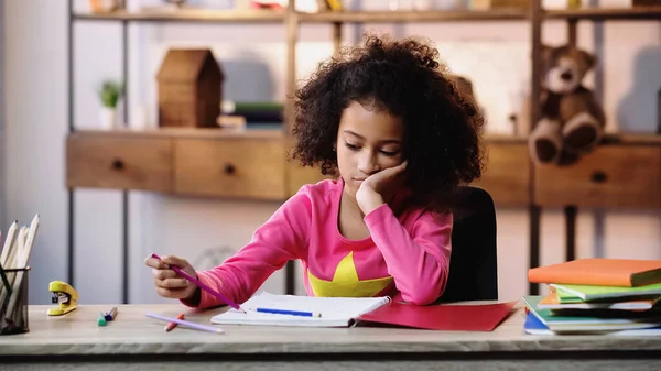 Bored african american schoolgirl looking at notebook while doing homework — Stockfoto