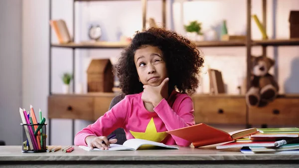 Thoughtful african american schoolgirl winking eye and doing homework — Fotografia de Stock