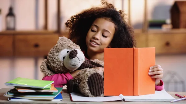 Happy african american girl reading book to teddy bear — Stockfoto