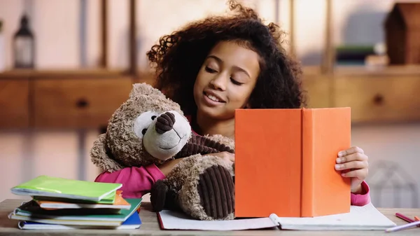 Happy african american girl reading book to teddy bear — стоковое фото