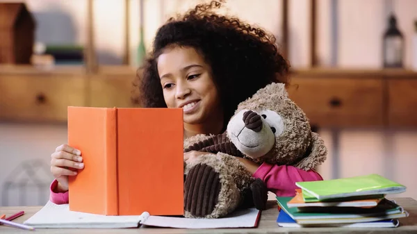 Happy african american schoolkid reading book to teddy bear — Stockfoto