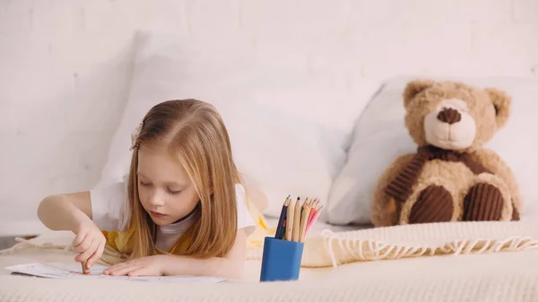 Детский рисунок с цветными карандашами рядом с размытым плюшевым мишкой на кровати — стоковое фото