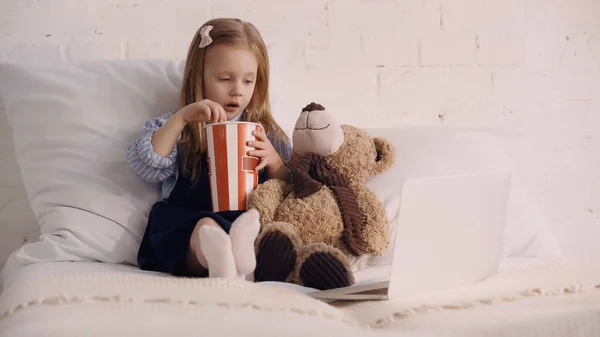 Child holding popcorn near teddy bear and laptop on bed — Stockfoto