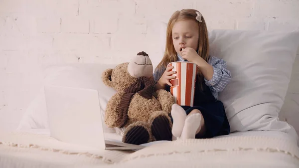 Kid eating popcorn near teddy bear and laptop on bed — Stock Photo