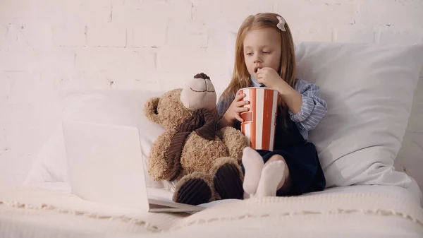 Niño comiendo palomitas de maíz cerca de oso de peluche y portátil en la cama - foto de stock