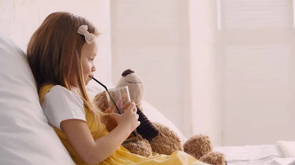 Vista lateral del niño bebiendo jugo cerca del oso de peluche en la cama - foto de stock