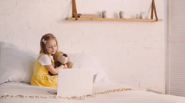 Lächelndes Kind umarmt Teddybär neben Laptop auf Bett — Stockfoto