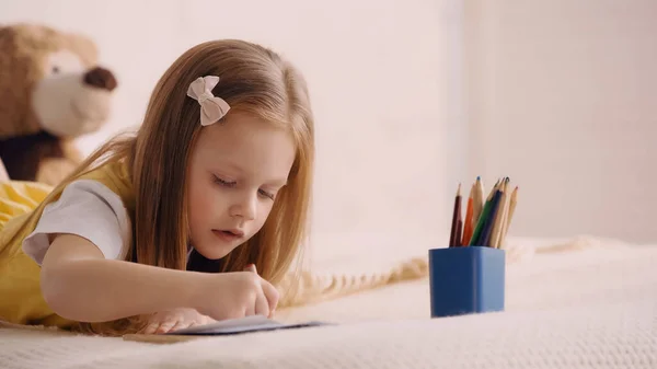 Child drawing on paper near color pencils on bed — Stock Photo