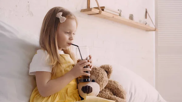 Chica bebiendo jugo con paja cerca de oso de peluche en la cama - foto de stock