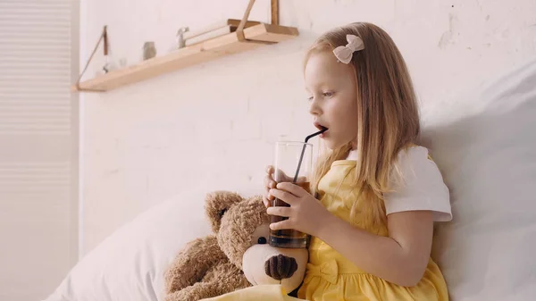 Kid holding glass of juice near teddy bear on bed — Stock Photo