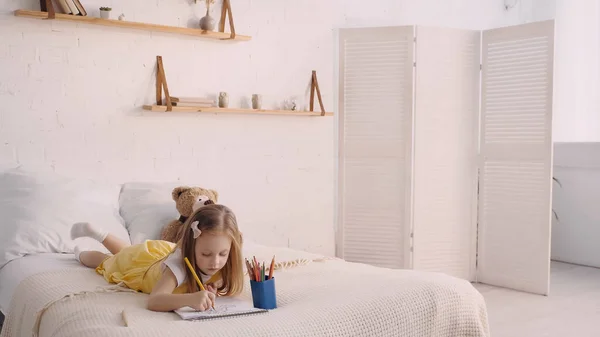 Criança desenho em papel perto de brinquedo macio na cama em casa — Fotografia de Stock