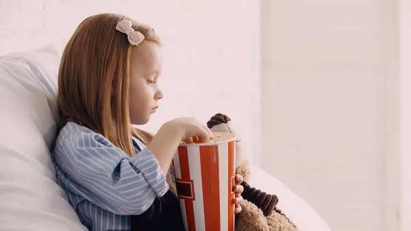 Side view of kid holding popcorn near soft toy on bed — Foto stock