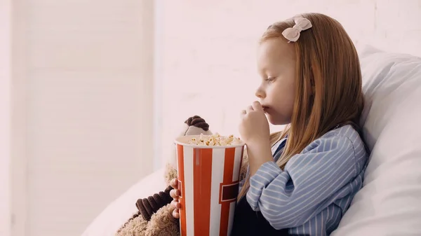 Side view of child eating popcorn near teddy bear on bed — Foto stock