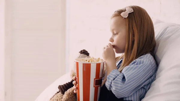 Side view of child eating popcorn near teddy bear on bed — Stock Photo
