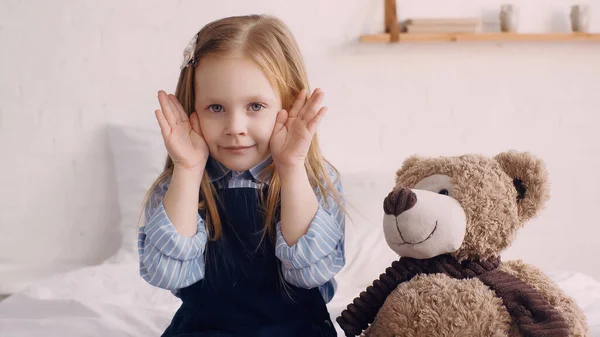 Child holding hands near face and looking at camera near soft toy on bed — Stock Photo