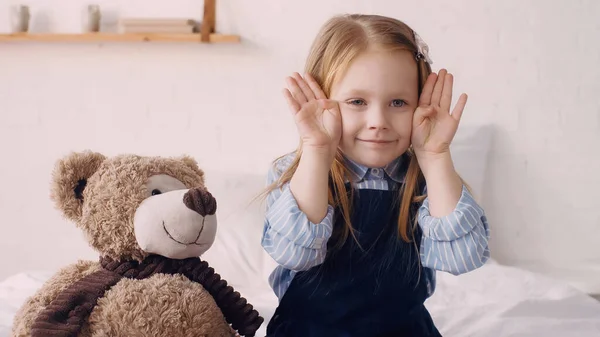 Smiling child holding hands near face and teddy bear on bed — Stock Photo