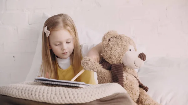 Kind zeichnet auf Skizzenbuch neben Teddybär auf Bett — Stockfoto