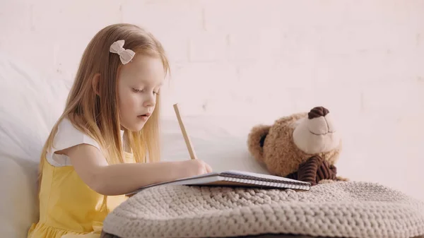 Preschooler kid drawing on paper near teddy bear and blanket at home — Stock Photo