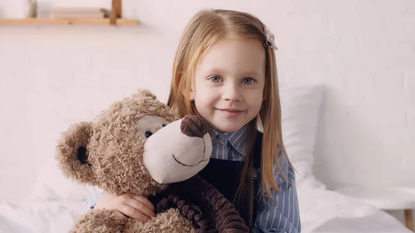 Positive kid hugging teddy bear and looking at camera in bedroom — Foto stock