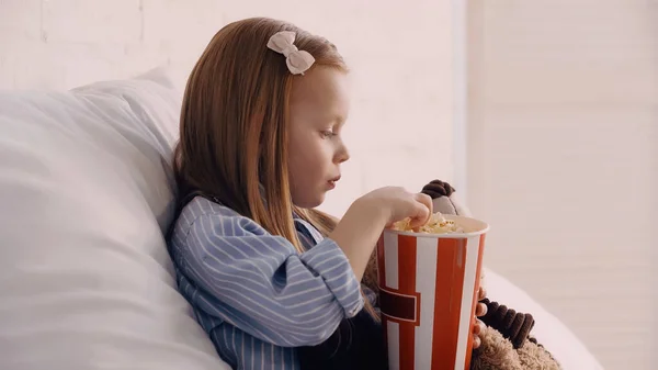 Side view of kid holding popcorn near soft toy in bedroom — Foto stock