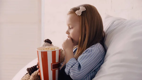 Side view of kid eating popcorn near toy on bed — Stock Photo