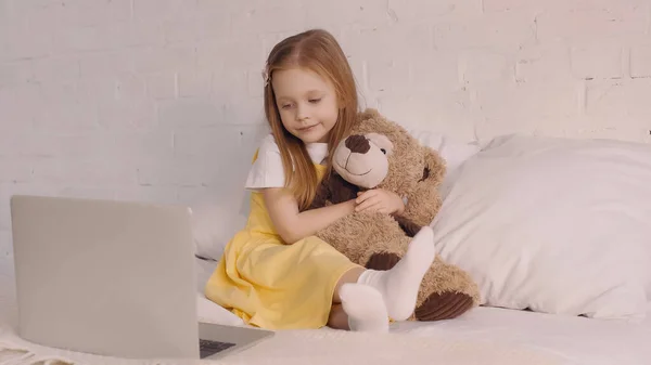 Child hugging teddy bear while looking at laptop in bedroom — Stock Photo