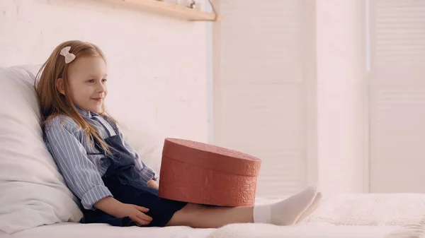 Enfant souriant assis sur le lit près de la boîte cadeau dans la chambre — Photo de stock