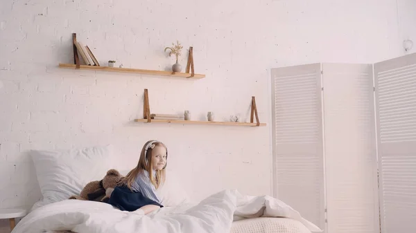 Positive kid in headphones looking at camera near soft toy in bedroom — Fotografia de Stock