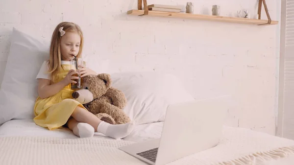 Child drinking juice near soft toy and laptop on bed — Stock Photo