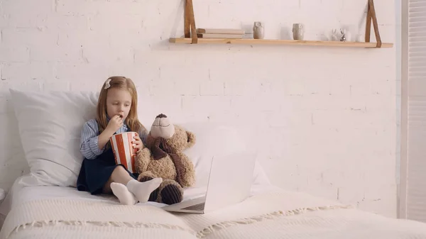 Kind isst leckeres Popcorn in der Nähe von Kuscheltier und Laptop im Schlafzimmer — Stockfoto