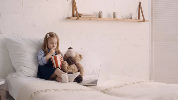 Kid eating popcorn while watching cartoons on laptop on bed — Stock Photo