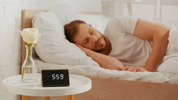 Rose and clock on bedside table near blurred man sleeping on bed — Foto stock