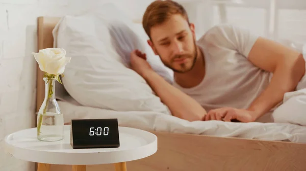 Clock and flower on bedside table near blurred man on bed at home — стоковое фото