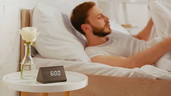 Clock and rose on bedside table near blurred man waking up on bed — Stock Photo