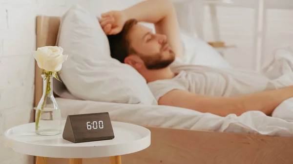 Rose in vase and clock on bedside table near blurred man lying on bed — Stock Photo