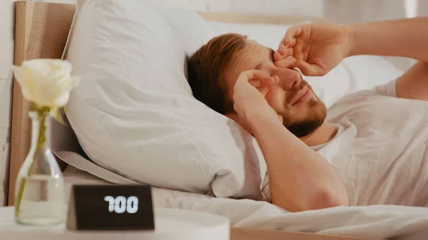 Schläfriger Mann reibt sich die Augen bei verschwommener Uhr auf dem Nachttisch zu Hause — Stockfoto