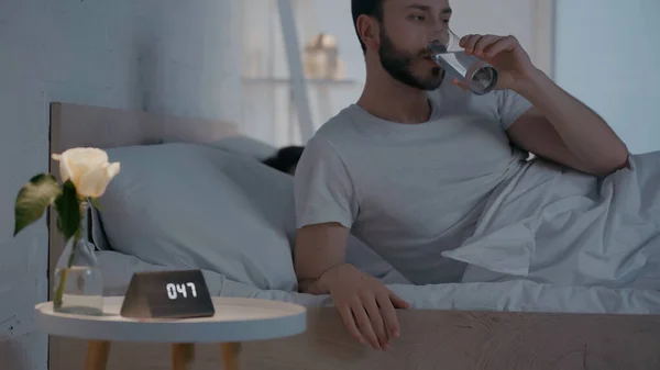 Man drinking water near clock on bedside table at night — Stock Photo