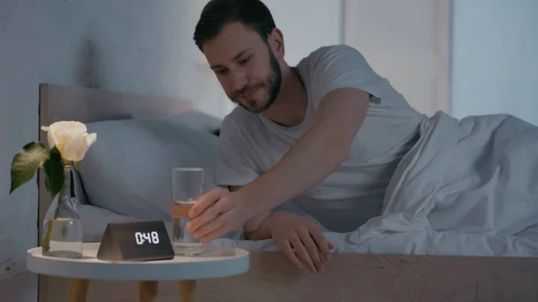 Young man taking glass of water near clock and plant on bedside table — Fotografia de Stock