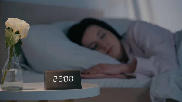 Clock and flower on bedside table near blurred woman sleeping on bed at night — Foto stock