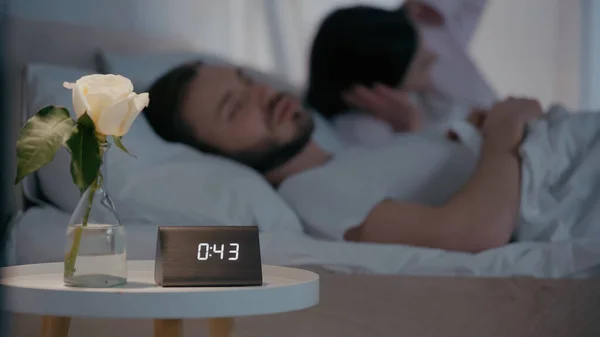 Flower and clock on bedside table near blurred man sleeping near wife at night — Stock Photo