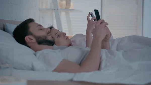 Young woman using smartphone near husband on bed at home — стоковое фото
