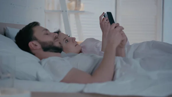 Mujer sonriente usando teléfono inteligente cerca de marido borroso en la cama - foto de stock