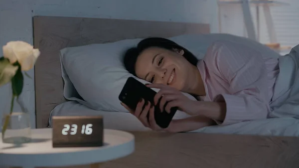 Cheerful woman using smartphone near blurred clock and plant in bedroom at night — Stock Photo